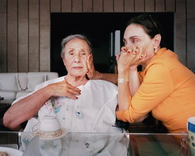 two woman in an interior seated at a glass table; elderly woman wearing white holds her hand to her chest and a younger woman wearing orange seated to the older woman's left holds her other hand to her face