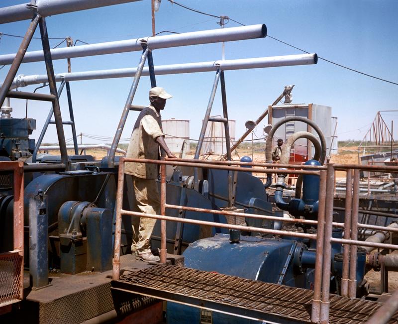 Color image of a man standing among machinery; two parallel silver pipes run over his head